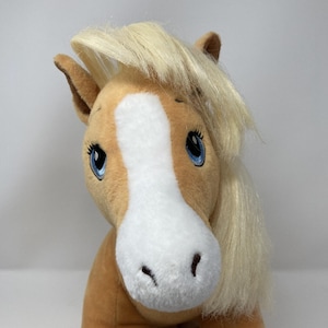 May include: A plush horse toy with tan fur, a white blaze on its face, and long, straight, pale blonde synthetic hair for a mane. The horse features large, embroidered blue eyes with black eyelashes and dark brown nostrils.