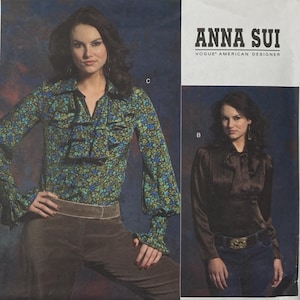 May include: A Vogue American Designer sewing pattern envelope by Anna Sui featuring two blouse designs. View C is a long-sleeved floral print blouse with ruffles, and View B is a solid brown satin blouse with a bow tie neck detail.