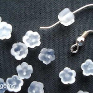 May include: A close-up of a group of clear plastic flower-shaped earring backs. The backs are scattered on a black surface. One earring back is attached to a silver earring hook.