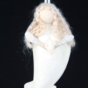 May include: A white, shell-shaped ornament with a white, glittery finish. The ornament is decorated with a white, felt-like figure with long, blonde hair. The figure is holding a seashell in each hand.