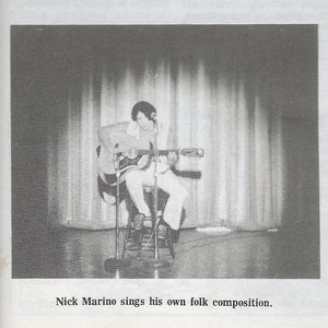 May include: A black and white photo of a person sitting on a stool and playing an acoustic guitar. The person is wearing a collared shirt and pants. The photo is taken from a slightly low angle. The caption below the photo reads "Nick Marino sings his own folk composition."