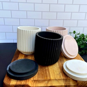 May include: A selection of concrete planters in cream, black, and blush pink. Each cylindrical planter has vertical ribbed detailing and a matching lid. The planters are displayed on a wooden chopping board.