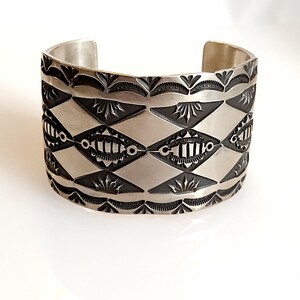 May include: A silver cuff bracelet with a black and white geometric design. The bracelet has a repeating pattern of diamonds and stylized eyes.