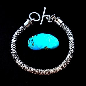May include: A silver chain bracelet with a toggle clasp. The bracelet is made of a woven chain pattern. A turquoise stone is shown next to the bracelet.