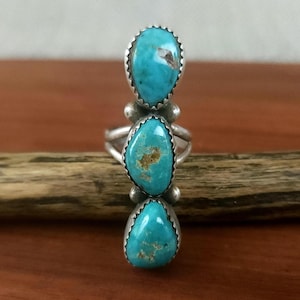May include: A silver ring featuring three vertically aligned, teardrop-shaped turquoise stones. Each stone is set in a serrated silver bezel, with small silver spheres placed between the stones. The band is split into two thin silver wires.