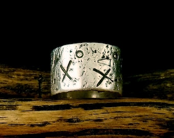 May include: A silver ring with a textured surface and engraved symbols. The ring is on a wooden surface.