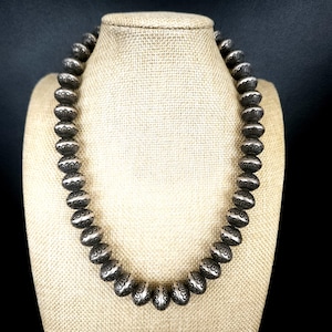 May include: A silver beaded necklace displayed on a mannequin. The necklace features large, round beads with etched floral patterns. The beads are strung together to form a complete necklace. The background is a dark color.