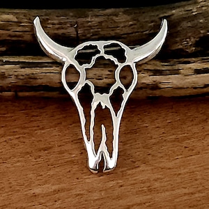 May include: A silver-colored metal brooch in the shape of a bull skull. The brooch features a detailed cutout design of the skull's features, including horns and facial structure. The brooch is displayed on a wooden surface.