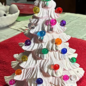 May include: A white ceramic Christmas tree with colorful, round lights. The tree has a textured surface and is decorated with green, red, yellow, blue, orange, and purple lights. The tree is on a red surface.