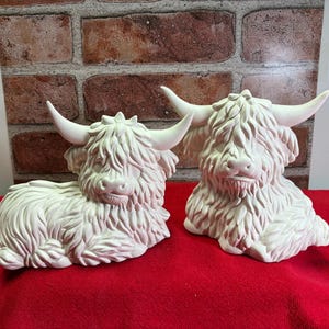 May include: Two unpainted ceramic figurines of Highland cattle, each featuring long, shaggy fur textures, curved horns, and detailed facial features. The figures are matte white, with one cow depicted in a lying position and the other in a sitting pose.