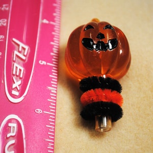 May include: Orange pumpkin-shaped glass ornament with a black painted face and a black and red pipe cleaner stem. The ornament is next to a pink ruler with the word "Flexi" printed on it.