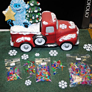 May include: A red toy truck with a white snowy roof, carrying a blue yeti holding a gold star and a green Christmas tree with colorful lights. Three bags of colorful Christmas lights are on a green surface with white snowflakes.