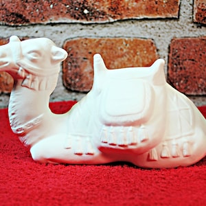 May include: White ceramic camel figurine with a saddle and tassels. The camel is lying down with its head turned to the side.