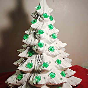 May include: A white ceramic Christmas tree with a green star on top and green shamrock-shaped ornaments. The tree is sitting on a red surface.