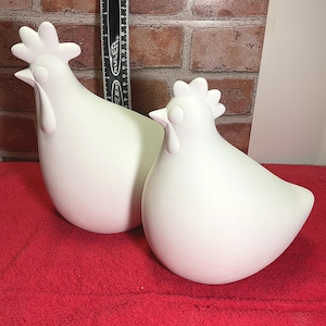 May include: Two white ceramic chicken figurines. The chickens are standing on a red surface.