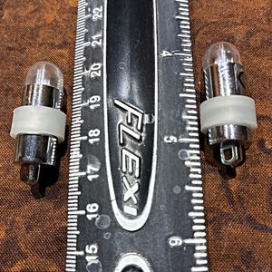 May include: Two small, clear light bulbs with silver bases and a white rubber ring. A black ruler with white markings is next to the bulbs. The bulbs are approximately 0.75 inches in length.