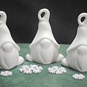 May include: Three white ceramic gnome ornaments with a rounded hat and a beard. The gnomes are sitting on a green surface with white snowflakes.