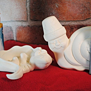 May include: Two white ceramic figurines of snails, one with a hat, on a red surface. A bottle of Duncan Bisque-Stain acrylic paint in gold is in the background.