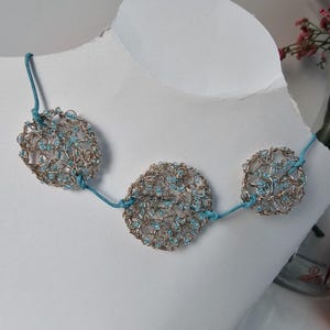 May include: A silver-toned wire necklace with three circular pendants. Each pendant is filled with small, light blue beads and connected by a turquoise cord. The necklace is displayed on a white mannequin.