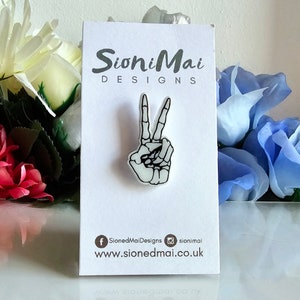 May include: White peace sign pin with a skeleton hand design. The pin is on a white card with the text "SioniMai Designs" and the website address "www.sionedmai.co.uk".