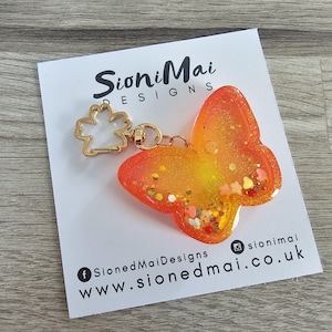 May include: A butterfly-shaped keyring with a gold-coloured clasp and a clear body filled with glitter and small heart-shaped confetti. The butterfly is orange and yellow. The keyring is on a white card with the text "SioniMai Designs".