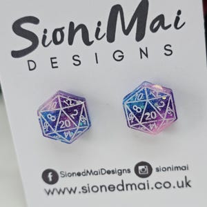 Puede incluir: Un par de pendientes hexagonales con forma de dados. Cada pendiente es un degradado de morado, azul y rosa con números blancos. Los pendientes están diseñados con un tema de galaxia.