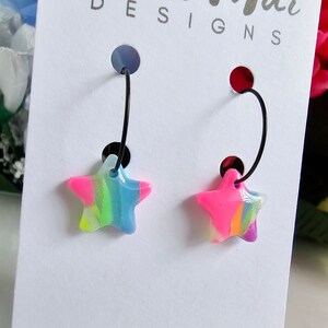 May include: A pair of black hoop earrings with colourful star-shaped charms. The stars have a pink, blue, green, and yellow swirl pattern.