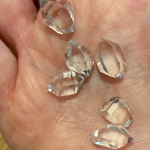 Peut inclure: Six pierres précieuses en cristal clair et facetté sont présentées dans une main. Les cristaux ont de multiples facettes, captant la lumière et créant un effet scintillant. Les pierres précieuses sont de forme irrégulière, avec des angles vifs et un aspect transparent.