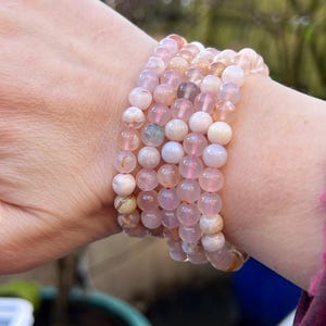Puede incluir: Varios brazaletes de cuentas en tonos rosa, melocotón y crema apilados en una muñeca. Los brazaletes están hechos de piedras redondas y pulidas. El fondo está borroso, con toques de follaje verde y una maceta azul.