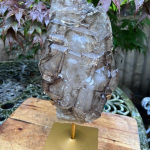 May include: A large, smoky quartz crystal cluster, mounted on a gold-colored metal stand. The crystal has a complex, geometric structure with varying shades of brown and gray. The stand has a square base and a cylindrical support.