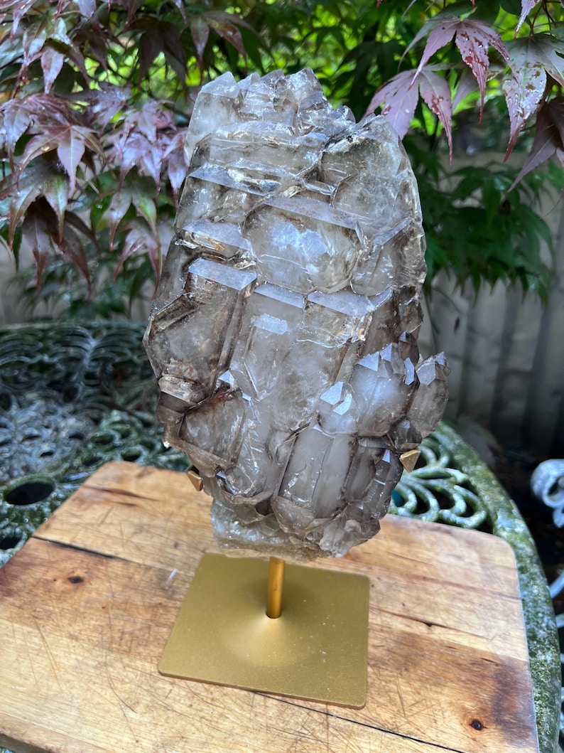 May include: A large, smoky quartz crystal cluster, mounted on a gold-colored metal stand. The crystal has a complex, geometric structure with varying shades of brown and gray. The stand has a square base and a cylindrical support.