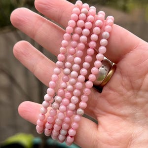 May include: A strand of pink gemstone beads, likely rose quartz, strung together to form a bracelet. The beads are small and round, and the bracelet is shown in a hand.