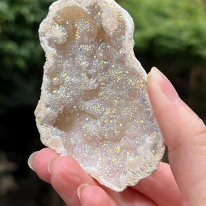 May include: A natural, irregularly shaped geode with a rough, beige exterior. The interior is filled with sparkling, iridescent crystals in shades of white, silver, and hints of rainbow colours. The geode is held in a hand.