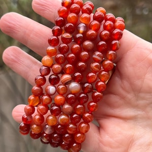 May include: A close-up of a stack of red carnelian gemstone beads. The beads are round and have a smooth, polished finish. The carnelian beads are a deep red color with white and orange stripes.