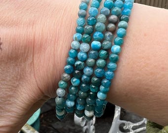 May include: Several beaded bracelets in shades of teal, turquoise, and grey. The bracelets are made of small, round beads and are worn on a wrist. The background is blurred, with hints of greenery and a white metal chair.