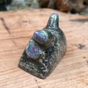 May include: A small, abstract sculpture with a dark green and gray mottled appearance. The sculpture has a rounded top and a smaller, rounded protrusion. The top and protrusion have iridescent purple and blue hues.