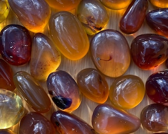 Puede incluir: Una colección de piedras pulidas y translúcidas en varios tonos ámbar, miel y marrón rojizo intenso. Estos guijarros suaves y redondeados exhiben variaciones naturales en color, claridad e inclusiones internas, creando un efecto cálido y brillante.