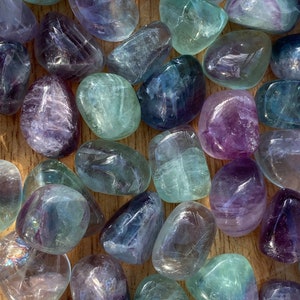 Puede incluir: Una colección de piedras de fluorita pulidas en tonos morados, azules y verdes. Las piedras pulidas tienen una forma lisa y redondeada y diversos grados de translucidez. Las piedras están dispuestas sobre una superficie de madera marrón claro.