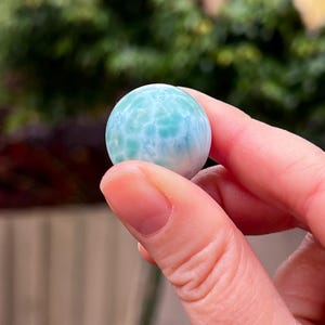 May include: A round, polished gemstone sphere, primarily light blue with white marbling. The stone is held between fingers, showcasing its smooth surface and intricate patterns. The background is blurred, with green foliage.