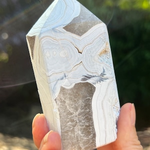 May include: White agate crystal point with a swirling pattern of white and gray bands. The crystal is polished and has a smooth, glassy surface.