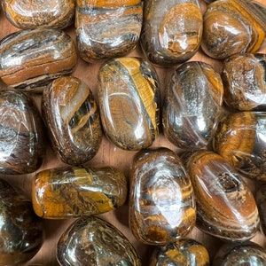 Könnte beinhalten: Eine Sammlung von polierten Tigeraugen-Steinen. Die Steine sind eine Mischung aus Braun, Gold und Schwarz mit einem glänzenden, irisierenden Finish.