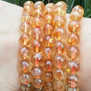 May include: Five bracelets composed of translucent, amber-coloured, spherical beads. Each bead is approximately 1.3 cm in diameter and strung on a red elastic cord. The bracelets are stacked on a wrist, with a green background.