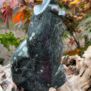 May include: A dark green carved stone sculpture of a cat. The cat sculpture has a textured surface with light green and pink accents. The sculpture is sitting on a piece of driftwood, with colorful fall foliage in the background.