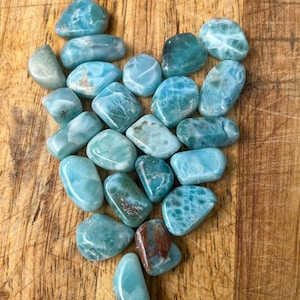 May include: A collection of blue and green tumbled stones arranged in a heart shape on a wooden surface. The stones have a marbled pattern and are smooth and polished.