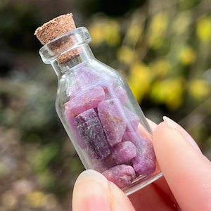 May include: A small glass bottle with a cork stopper filled with pink and purple gemstone chips.