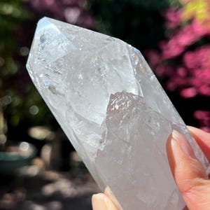 Könnte beinhalten: Ein klarer Quarz-Kristallpunkt, gehalten in einer Hand, mit einer facettierten, polierten Oberfläche. Der Kristall ist transparent und lässt Licht durch. Der Hintergrund ist verschwommen, mit grünem und rosa Laub.