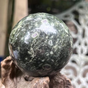 May include: A polished spherical stone featuring a deep forest green base color with mottled light green and white inclusions, along with small, distinct dark red spots scattered across the smooth, reflective surface of the orb.