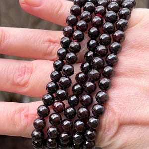 May include: A strand of dark red garnet beads, with a shiny, polished finish. The beads are round and uniform in size, and they are strung on a clear elastic cord.