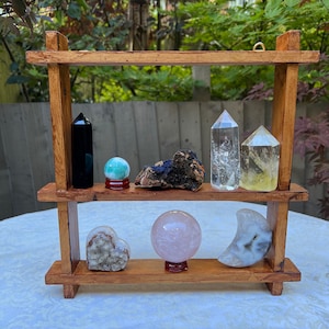 Op de afbeelding: Een houten plank met twee niveaus met diverse kristallen en edelstenen. De plank bevat een zwarte obsidiaantoren, een groene bol, een blauw en bruin mineraal, heldere kwartspunten, een gele kwartspunt, een hartvormig kristal, een roze bol en een maanvormige steen.