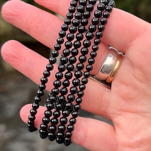 Puede incluir: Seis hilos de pulseras de cuentas negras se sostienen en una mano. Las cuentas, pequeñas y redondas, tienen un acabado brillante. Las pulseras están drapeadas sobre la palma, mostrando su color oscuro y textura. Se ve un anillo de oro y plata.